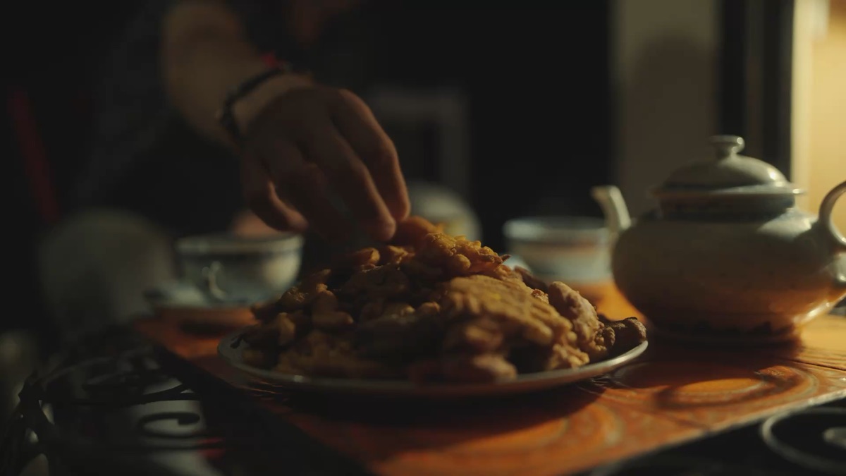 Film still from the Anju commercial — a hand reaching for fresh pakoras beside a teapot, warm lamplight