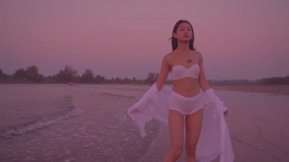 Still frame from the Pranisha music video — model walking toward camera on a pink sunset beach, white fabric in the wind